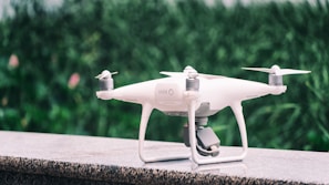 A white quadcopter drone is placed on a marble surface with a blurred background of green foliage. It features four propellers and a camera mounted on the underside.