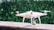 A white quadcopter drone is placed on a marble surface with a blurred background of green foliage. It features four propellers and a camera mounted on the underside.