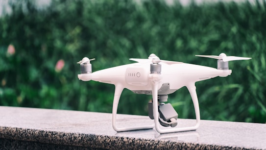 A white quadcopter drone is placed on a marble surface with a blurred background of green foliage. It features four propellers and a camera mounted on the underside.