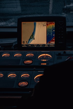 A control panel with illuminated gauges and a digital display showing a navigation map and sonar data. The display indicates speed, heading, and depth information. The ambient lighting is dim, emphasizing the glow from the screen and gauges.