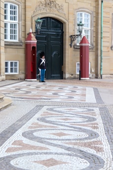 An ornate building entrance with a dark green door is flanked by two decorative red guard boxes. A ceremonial guard dressed in a traditional uniform with a black fur hat stands by the door. The pavement features intricate patterns made with small tiles in white, beige, and brown colors.