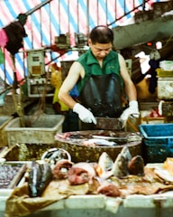 A market vendor wearing a green sleeveless shirt and black apron stands behind a table, chopping fish with a cleaver. Various fish parts and seafood are spread out on the table in front of him. The background features a striped tarpaulin and market goods stacked around.