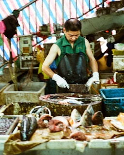 A market vendor wearing a green sleeveless shirt and black apron stands behind a table, chopping fish with a cleaver. Various fish parts and seafood are spread out on the table in front of him. The background features a striped tarpaulin and market goods stacked around.