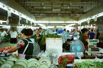 A bustling indoor market scene with numerous people engaged in selling or purchasing fresh produce. Stalls are filled with vegetables and fruits, such as melons and tomatoes. Electric fans are placed around to provide ventilation, and scales are used for weighing the goods.