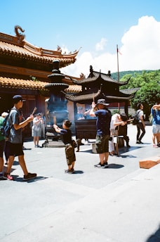 People are gathered in front of a traditional Chinese temple with ornate roofs, participating in a cultural or religious activities involving incense. The area is sunlit, casting clear shadows, and the sky is bright and clear.
