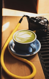 A blue cup filled with matcha green tea sits on a wooden surface, accompanied by a camera with a yellow strap. Sunlight casts shadows on the wood, creating a warm atmosphere. The latte art on the tea forms a leaf pattern.