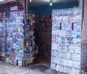 A small, bustling kiosk filled with displays of CDs or DVDs in transparent plastic cases. The walls and shelves are densely covered with cover art, displaying a wide variety of movies or music albums. The kiosk is situated in an outdoor setting, and a single lightbulb hangs from the ceiling, providing illumination. A partially visible person is sitting inside, possibly organizing or attending to the store.