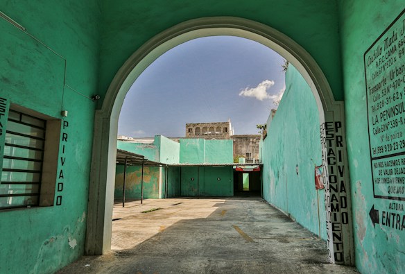 The image features an arched entrance leading into an open courtyard with turquoise walls. Signs with Spanish text are visible, indicating it is a private parking area. The courtyard is empty, with a shadow on the concrete floor and a partially visible building in the background beneath a clear sky.
