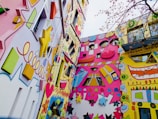 A vividly painted building exterior featuring a mix of geometric shapes and stylized faces. The design includes stars, hearts, flowers, and spirals, creating a lively and whimsical atmosphere. The facades are adorned with bright colors like pink, yellow, and blue, and various elements project outward in a playful manner. There is a tree with thin branches visible in the foreground.