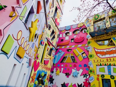 A vividly painted building exterior featuring a mix of geometric shapes and stylized faces. The design includes stars, hearts, flowers, and spirals, creating a lively and whimsical atmosphere. The facades are adorned with bright colors like pink, yellow, and blue, and various elements project outward in a playful manner. There is a tree with thin branches visible in the foreground.