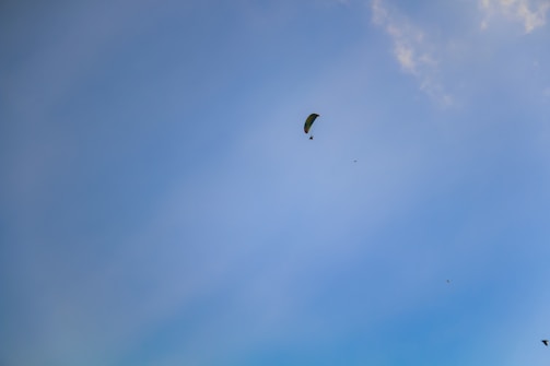 A paraglider is visible in the sky, soaring amidst a few scattered clouds. The sky is predominantly clear, with a few birds flying nearby.