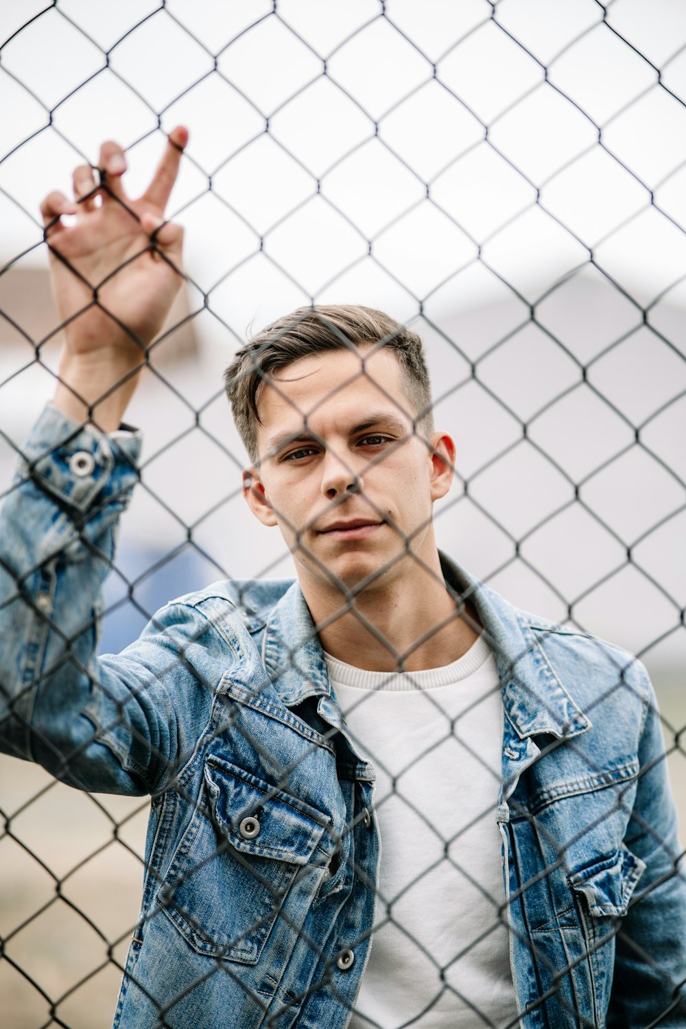 photo of man wearing of white crew-neck shirt and blue denim faded jacket behind of cyclone fence