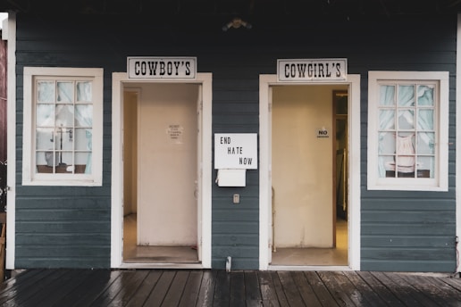 A rustic wooden building features two doors labeled 'Cowboy's' and 'Cowgirl's' with white-framed windows. A sign between the doors reads 'End Hate Now'. The exterior is painted dark green with white trim, and the walkway is wooden.