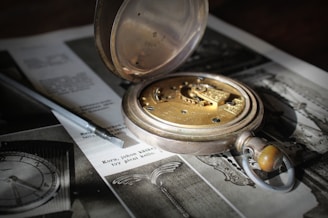 A vintage pocket watch with an open cover exposing its intricate golden gears and mechanisms. The watch is placed on a black and white printed page with an old photograph and some text. A screwdriver lies beside the watch, suggesting repair or assembly work.