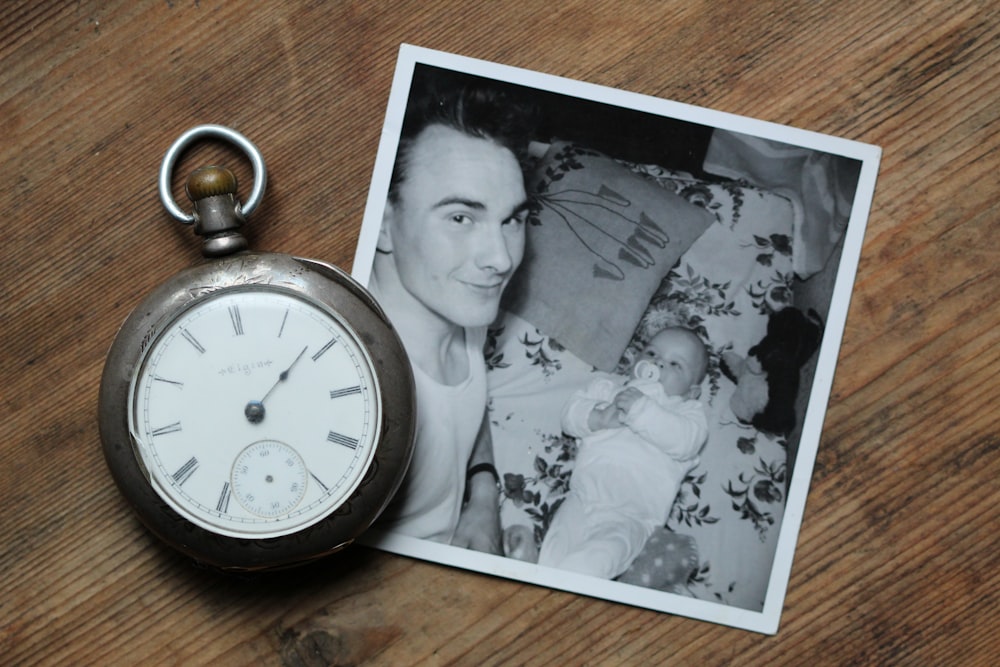 テーブルの上の父と赤ん坊のグレースケール写真の横にある灰色の懐中時計