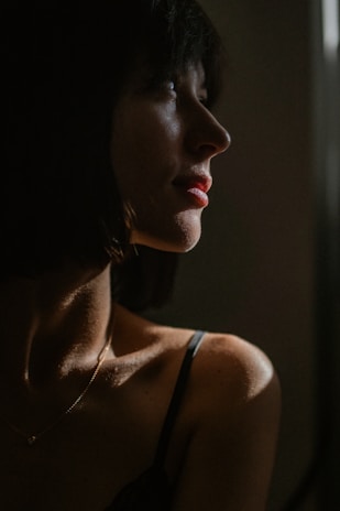 A person with short dark hair is seen in profile, illuminated by soft, dramatic lighting that casts shadows on their face and shoulder. The lighting highlights the contours of the face, emphasizing the person's features while creating a moody atmosphere.