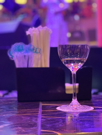 A clear wine glass stands elegantly on a reflective marble surface, partially filled with liquid. Behind the glass, a container holds a selection of white straws and blue swizzle sticks. The background is softly blurred with vibrant hues of purple, pink, and yellow, suggesting a lively, ambient setting.