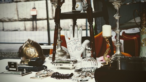 A collection of antique and vintage items displayed on a table, including a brass helmet, a palmistry hand model, various candlesticks, and a selection of small decorative objects. The scene gives a sense of eclectic charm and nostalgia.