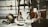A collection of antique and vintage items displayed on a table, including a brass helmet, a palmistry hand model, various candlesticks, and a selection of small decorative objects. The scene gives a sense of eclectic charm and nostalgia.