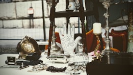 A collection of antique and vintage items displayed on a table, including a brass helmet, a palmistry hand model, various candlesticks, and a selection of small decorative objects. The scene gives a sense of eclectic charm and nostalgia.