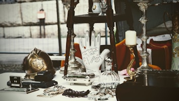 A collection of antique and vintage items displayed on a table, including a brass helmet, a palmistry hand model, various candlesticks, and a selection of small decorative objects. The scene gives a sense of eclectic charm and nostalgia.