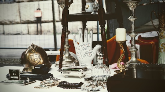 A collection of antique and vintage items displayed on a table, including a brass helmet, a palmistry hand model, various candlesticks, and a selection of small decorative objects. The scene gives a sense of eclectic charm and nostalgia.