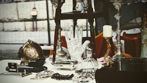 A collection of antique and vintage items displayed on a table, including a brass helmet, a palmistry hand model, various candlesticks, and a selection of small decorative objects. The scene gives a sense of eclectic charm and nostalgia.