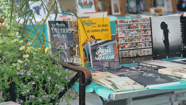A variety of books are displayed on a table, with colorful covers featuring titles in different languages. Some books are propped up, while others lie flat. In the foreground, there's a lush green plant with small purple flowers. The setting appears to be an outdoor book market.