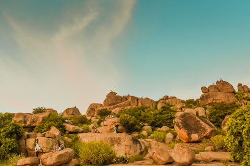 A rocky landscape features large boulders scattered across a lush, green environment. There are people sitting and climbing on the rocks. The sky is clear with a hint of clouds, creating a serene and natural setting.