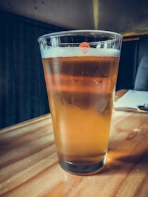 A pint glass is filled with amber-colored beer, topped with a frothy head. The glass rests on a wooden table with soft lighting creating a cozy ambiance. In the background, there are blurred elements of what appears to be a bar or cafe setting.