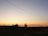 A tranquil rural landscape at sunset with a clear sky transitioning from yellow to blue. Power lines stretch across the sky, and there are a few trees silhouetted against the horizon. The ground is mostly dark, with a faint outline of hills in the background.