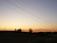 A tranquil rural landscape at sunset with a clear sky transitioning from yellow to blue. Power lines stretch across the sky, and there are a few trees silhouetted against the horizon. The ground is mostly dark, with a faint outline of hills in the background.