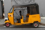 A yellow auto rickshaw is parked on the side of the road in front of a gray wooden door. The vehicle has a black canopy and a license number displayed on its side. The setting appears urban with a paved sidewalk.