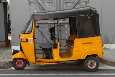 A yellow auto rickshaw is parked on the side of the road in front of a gray wooden door. The vehicle has a black canopy and a license number displayed on its side. The setting appears urban with a paved sidewalk.