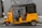 A yellow auto rickshaw is parked on the side of the road in front of a gray wooden door. The vehicle has a black canopy and a license number displayed on its side. The setting appears urban with a paved sidewalk.