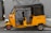 A yellow auto rickshaw is parked on the side of the road in front of a gray wooden door. The vehicle has a black canopy and a license number displayed on its side. The setting appears urban with a paved sidewalk.