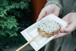 A rectangular white plate holds a skewer with a grilled rice cake topped with small white fish and herbs. Hands are holding the plate, and the background features green foliage.