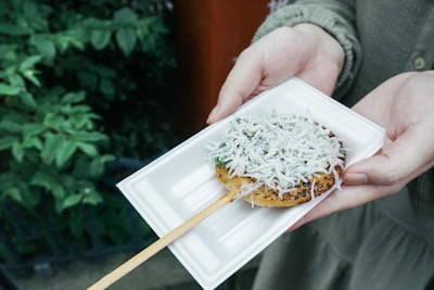 A rectangular white plate holds a skewer with a grilled rice cake topped with small white fish and herbs. Hands are holding the plate, and the background features green foliage.