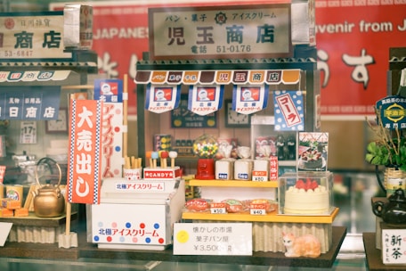 A colorful miniature display featuring a Japanese-themed shop with tiny, detailed items such as candies, a kettle, and a cake. The shop is adorned with various Japanese signs, banners, and a small model of a cat. A mix of traditional and playful elements gives it a nostalgic and charming atmosphere.