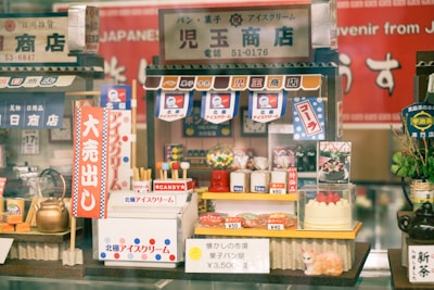 A colorful miniature display featuring a Japanese-themed shop with tiny, detailed items such as candies, a kettle, and a cake. The shop is adorned with various Japanese signs, banners, and a small model of a cat. A mix of traditional and playful elements gives it a nostalgic and charming atmosphere.