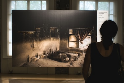 A woman stands in front of a large photographic display in an art gallery. The photograph depicts an abandoned room with dusty floors, cracked walls, and a window with tattered curtains. Debris and old objects are scattered around the room, creating a scene of neglect and decay. Natural light filters in through the windows of the gallery, casting a soft glow.