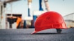 A bright red safety helmet is placed on a rough black surface in a construction site. The background is blurred but shows construction structures and cranes, creating an industrial atmosphere.