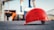 A bright red safety helmet is placed on a rough black surface in a construction site. The background is blurred but shows construction structures and cranes, creating an industrial atmosphere.