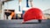 A bright red safety helmet is placed on a rough black surface in a construction site. The background is blurred but shows construction structures and cranes, creating an industrial atmosphere.