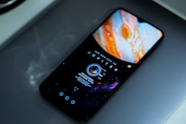 A smartphone displays an image of Jupiter on its screen. The phone rests on a metallic surface, surrounded by a soft, diffuse lighting that casts a faint reflection.