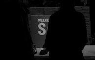 A dimly lit scene with people standing around a table. In the background, stacks of shoeboxes are visible. A prominent sign on the table reads 'WEEKEND SALE' in large letters. The colors are primarily dark with silhouettes of people.