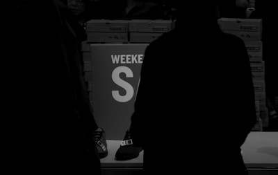 A dimly lit scene with people standing around a table. In the background, stacks of shoeboxes are visible. A prominent sign on the table reads 'WEEKEND SALE' in large letters. The colors are primarily dark with silhouettes of people.