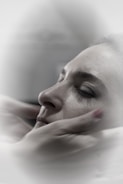 A close-up of a person with closed eyes, being gently cradled by hands which suggest a comforting or soothing gesture. The soft focus and muted colors create a calm, serene atmosphere.