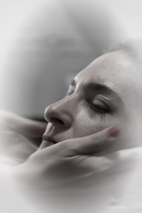 A close-up of a person with closed eyes, being gently cradled by hands which suggest a comforting or soothing gesture. The soft focus and muted colors create a calm, serene atmosphere.