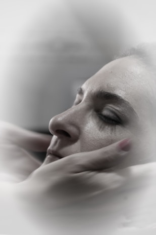 A close-up of a person with closed eyes, being gently cradled by hands which suggest a comforting or soothing gesture. The soft focus and muted colors create a calm, serene atmosphere.
